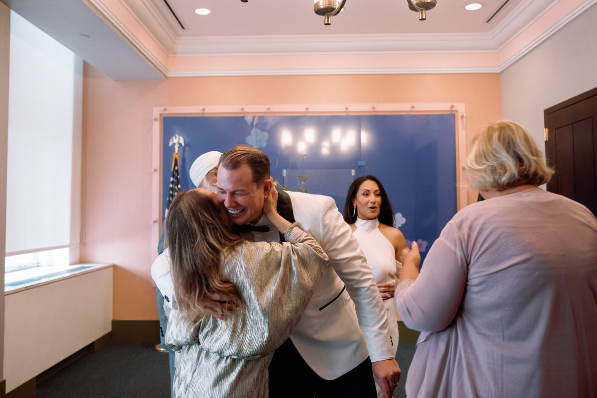 Groom Chris shares a joyful hug with a guest while family gathers around during the City Hall wedding celebration