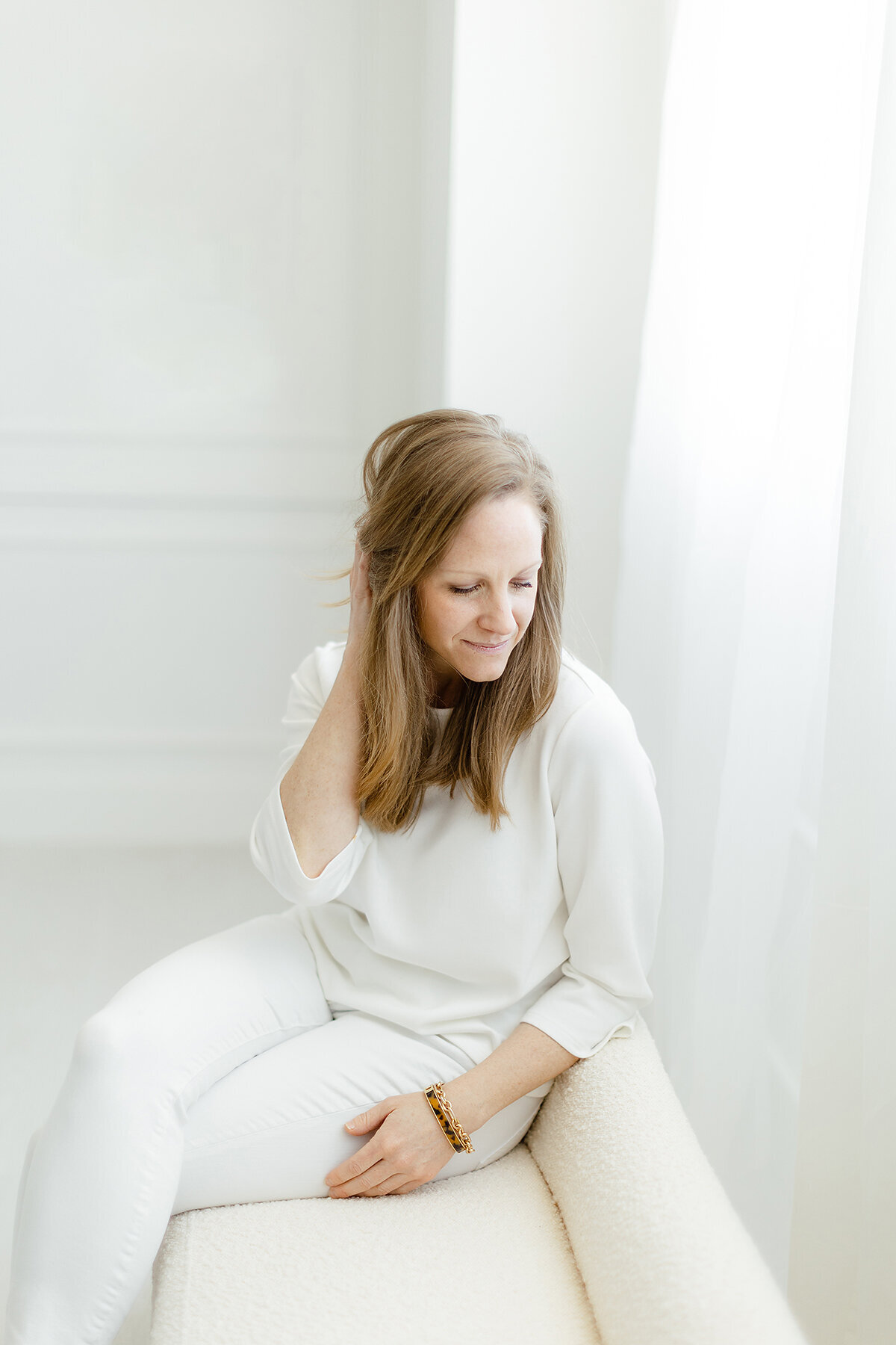 Light filled in studio professional headshot photo of a Dallas woman sitting down on a chair while she holds her hair behind her head and looks down.