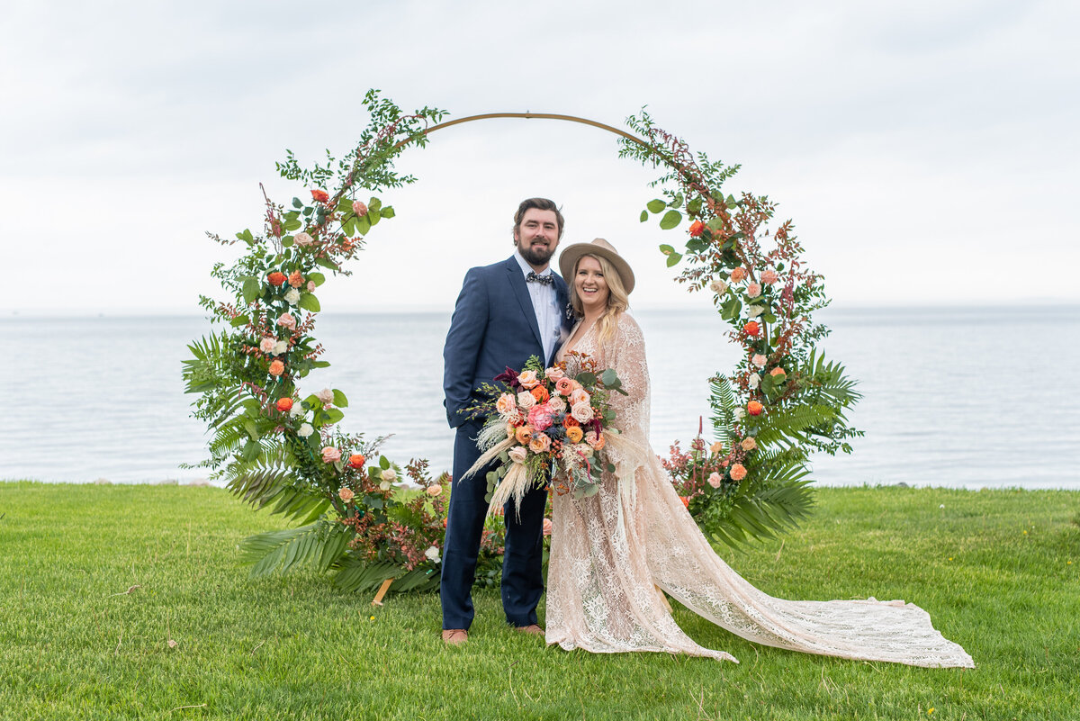 Boho styled bride and groom embrace and smile in front of lush floral circular arch