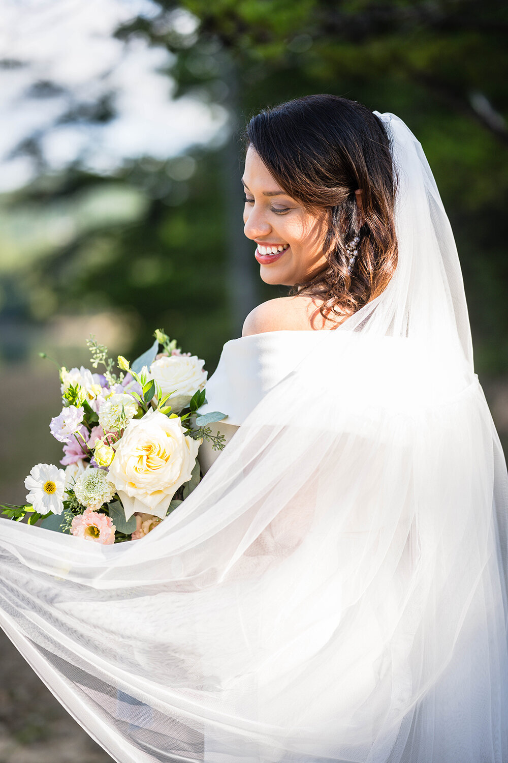 A marrier faces away from the photographer and looks past her shoulder at a bouquet of pastel flowers as her veil cascades from where it’s pinned on her head towards the bottom left corner of the frame.