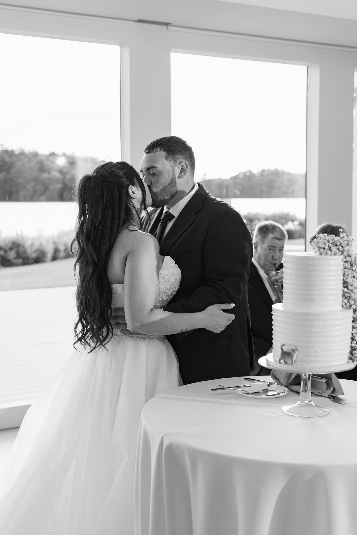 The Lakehouse Inn MA  | Kelsey Sheehan Photography Timeless Rhode Island Weddings | A newlywed couple shares a kiss beside a tiered wedding cake indoors. A guest watches in the background. The scene is set with large windows and natural light.