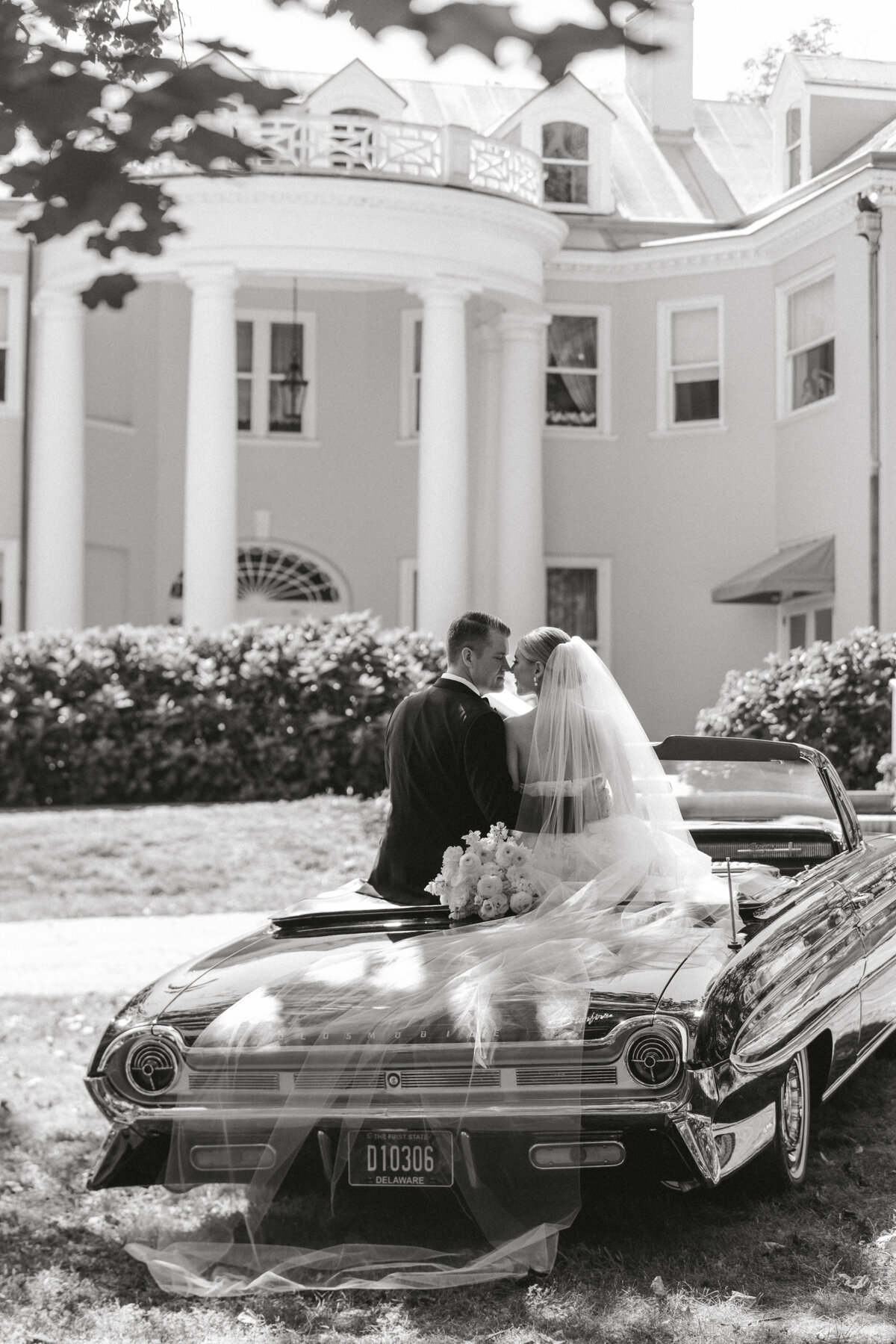 Bride and groom sit kissing on the back of a vintage car. Bellevue Hall is in the background.