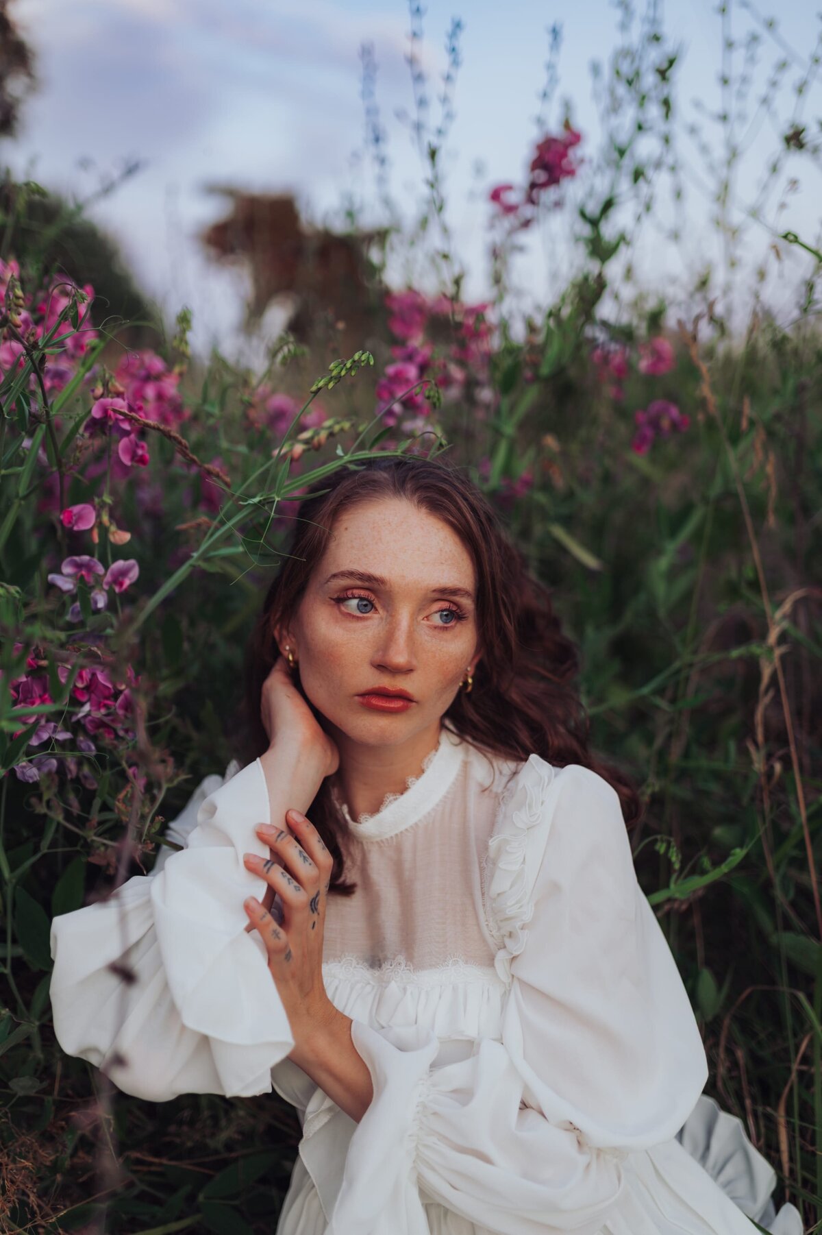 Young, red-headed woman sits in a flowy, white dress in a field of flowers while looking away from the camera and posing