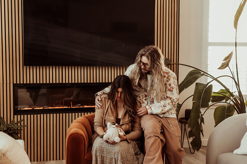 Mom and dad snuggling their newborn baby while sitting on a modern chair in their living room.