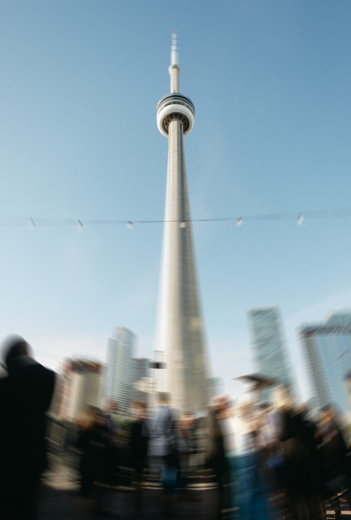 The CN Tower under a clear blue sky, with the blurred silhouettes of people in the foreground, captures a moment akin to the artistry of a destination wedding photographer.