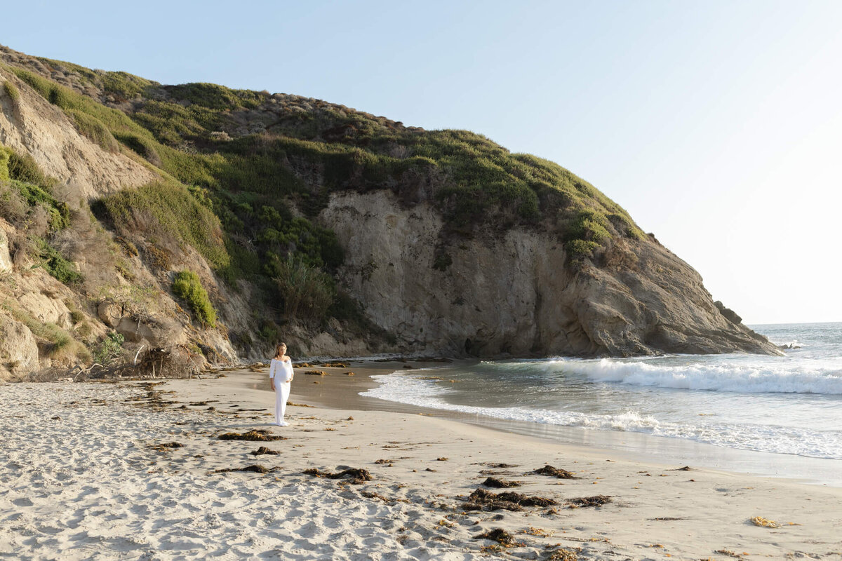 A person in white clothing stands on a sandy beach with gentle waves approaching the shore. In the background, there is a large, rocky cliff covered with green vegetation. The sky is clear and bright.