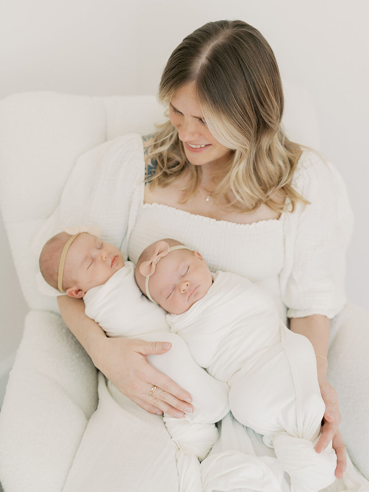 A mother lovingly cradles her newborn twins in her arms, holding them close to her chest. She gazes at them with warmth and tenderness as they rest peacefully. The soft natural lighting highlights the beauty of this intimate moment, capturing the deep bond between mother and babies.