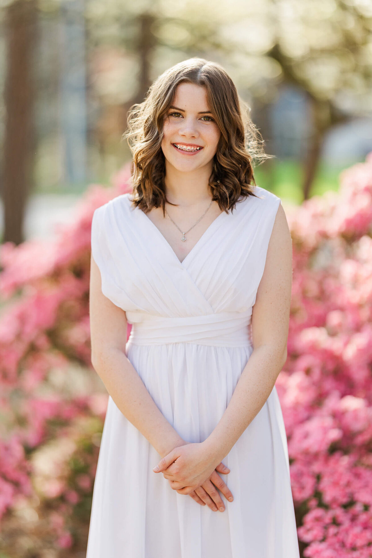 A young woman with wavy brown hair is wearing a white sleeveless dress, smiling brightly. She stands in front of blooming pink flowers, with blurred trees in the background—captured beautifully by a Fayetteville NC family photographer.