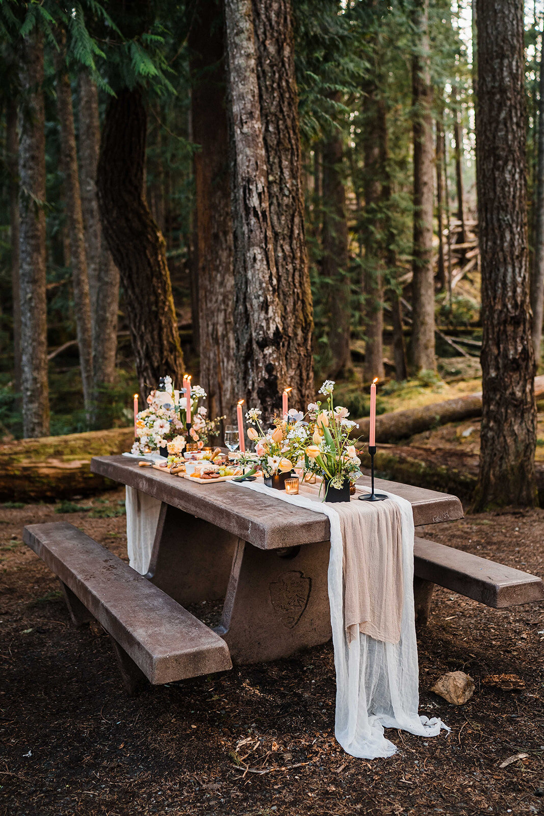 Picnic table in the woods decorated with gauzy runners, colorful flowers & candlesticks for elopement celebration!