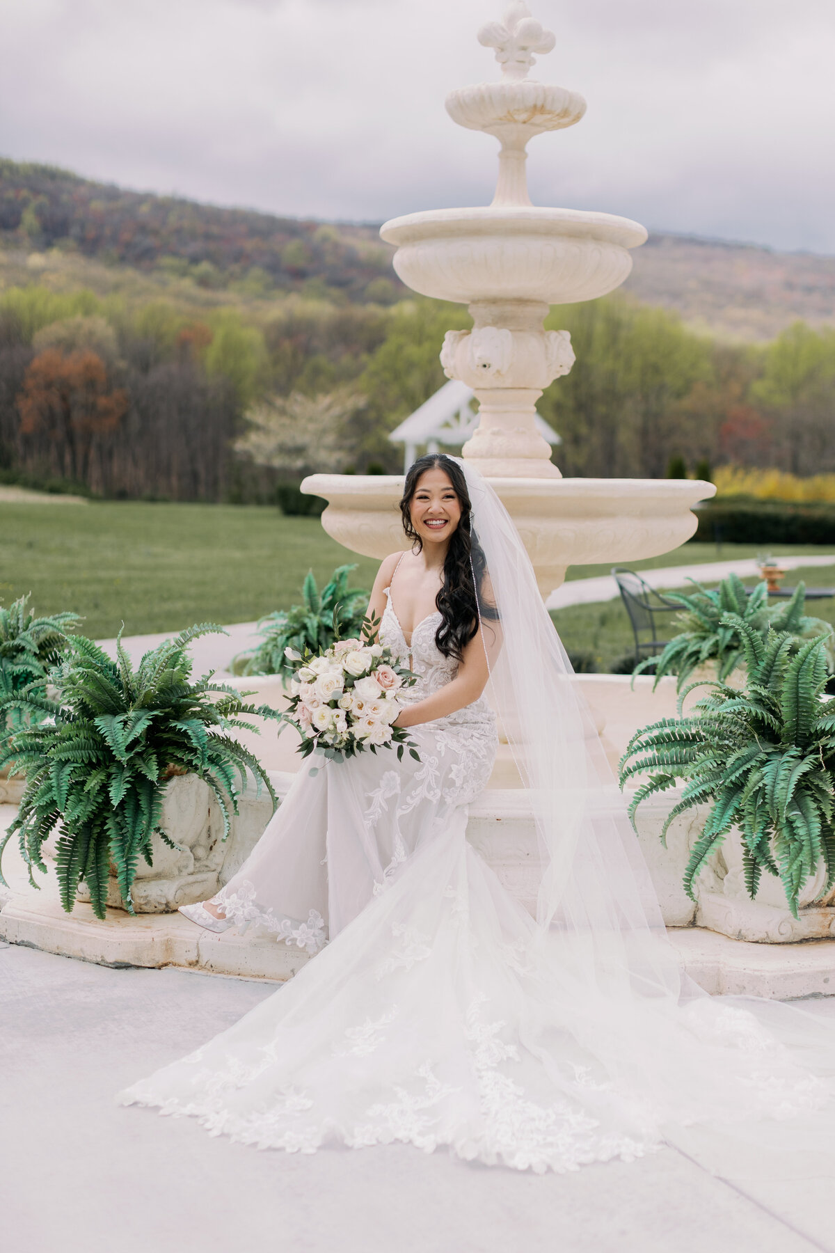Bride in lace gown holding bouquet by Flowers by Wendy Carol beside fountain at Springfield Manor.