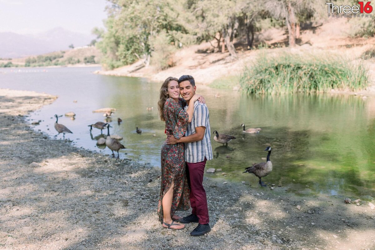 Engaged couple embrace on another next to a lake at Frank G Bonelli Park next to geese