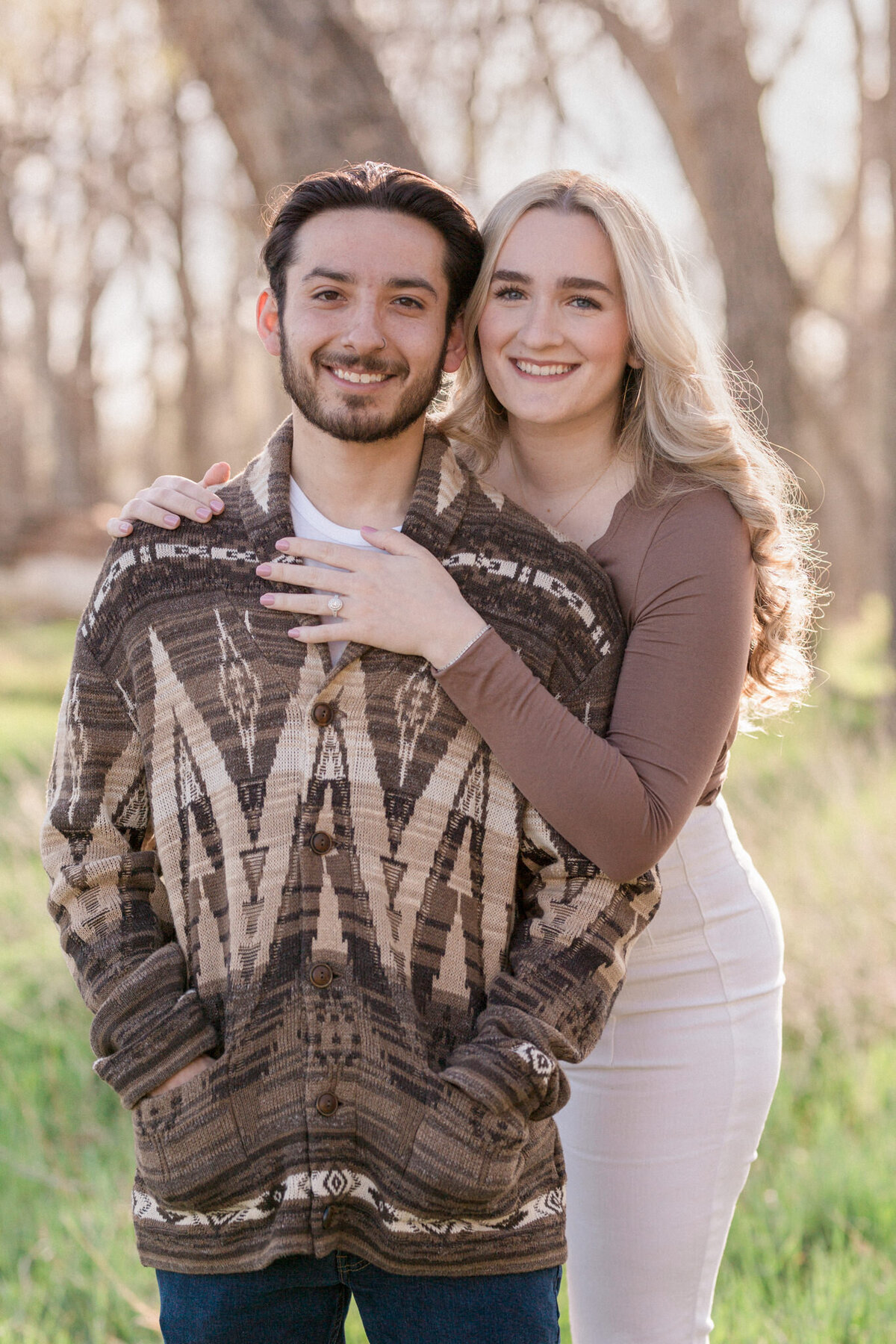 A man stands, hands in pockets with his new fiancee hugging him from behind, her hand on his chest to show off her engagement ring