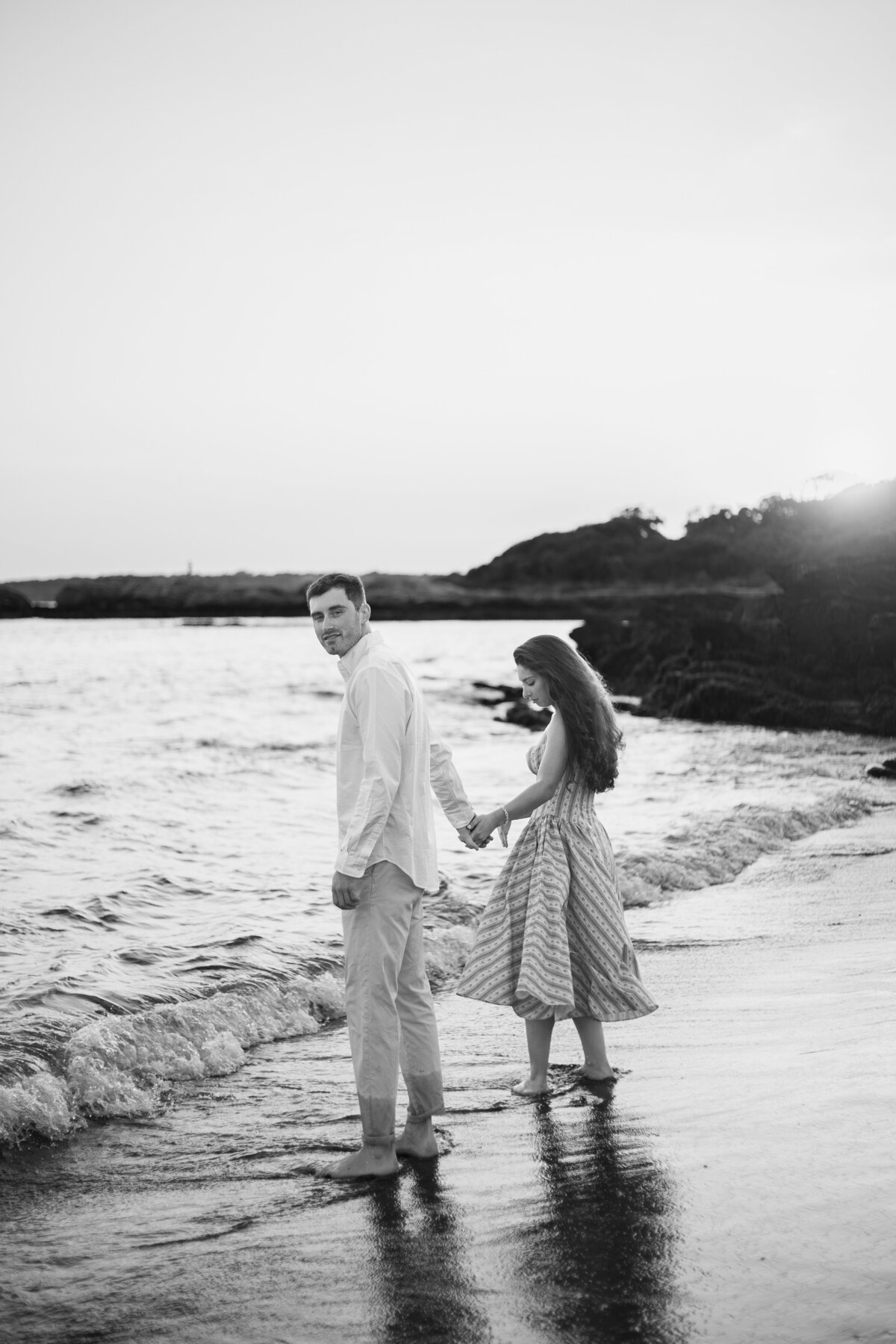 Engagement Photos Rhode Island | Kelsey Sheehan Photography Timeless Rhode Island Weddings | A couple holds hands, walking barefoot on a beach at sunset. The scene is serene and romantic, with gentle waves and a rocky shoreline in the background.