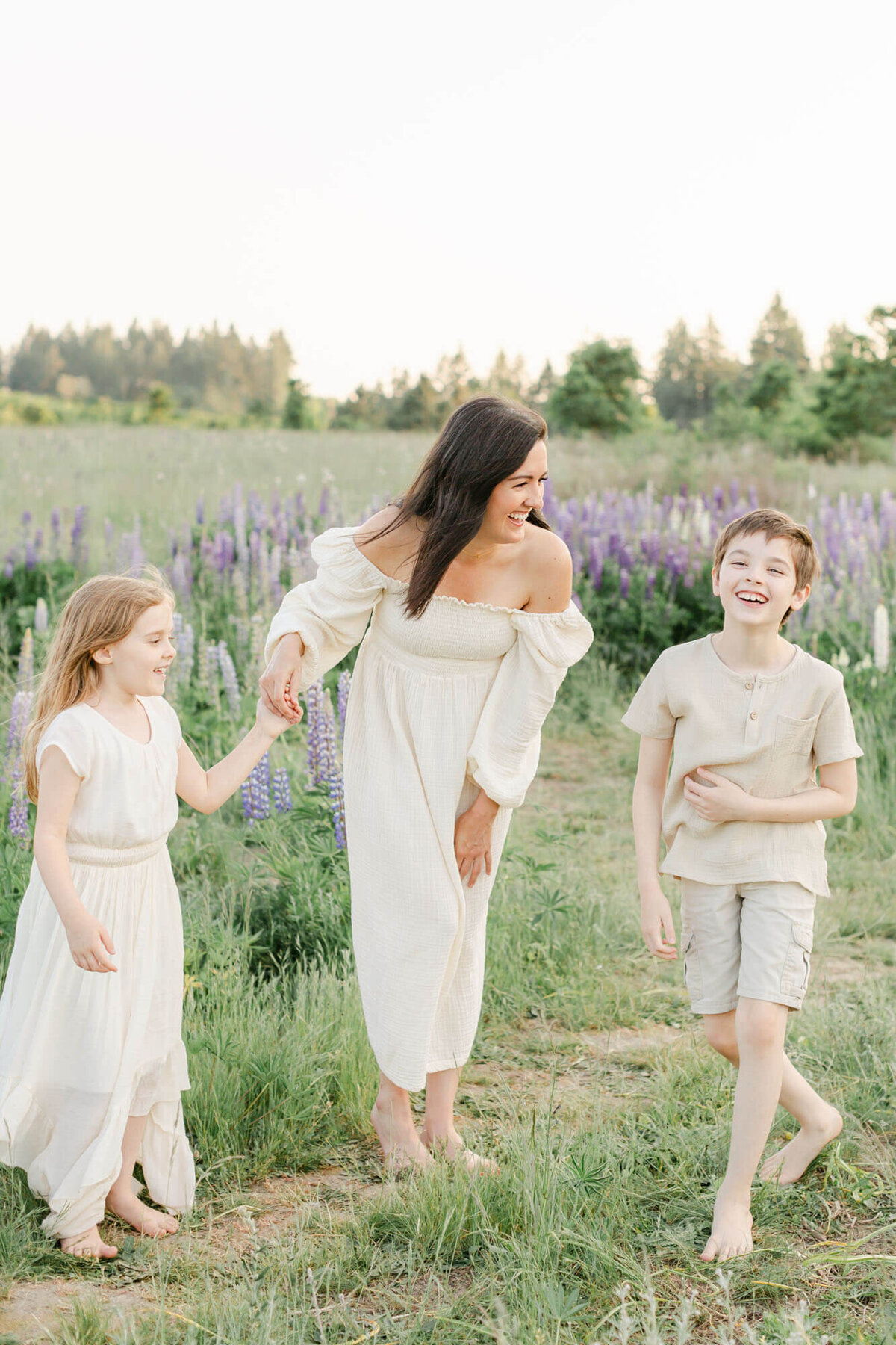 Gorgeous Mama in cream Nothing Fits But dress and long dark hair is out in a field of blue and purple wildflowers with her two children. They are all laughing together. Her kids are both dressed in light neutral tones and colors. She is looking at her son who is standing to her left and her daughter is holding her hand on her right.