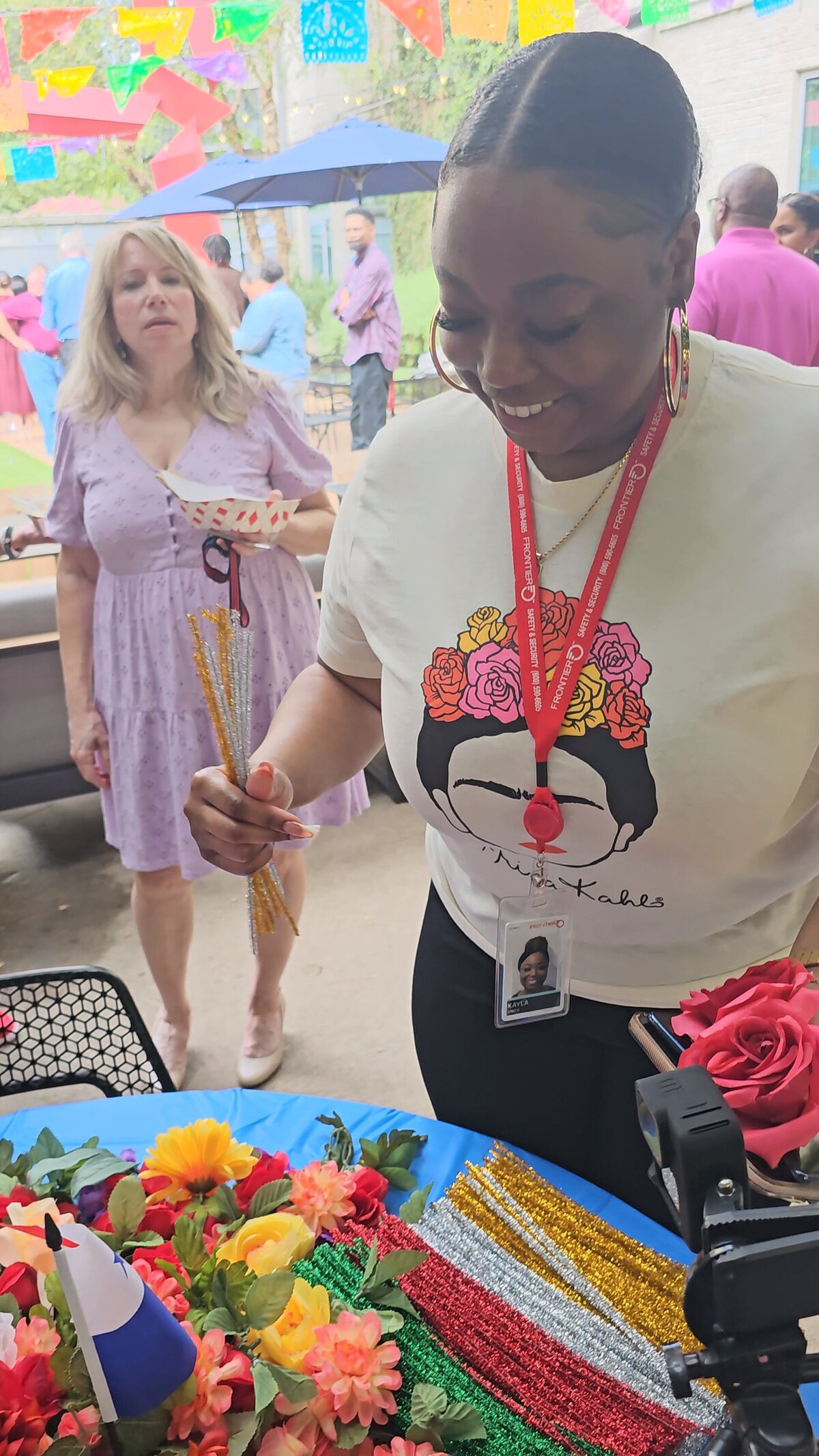 The magic of artistry as a woman, dressed in a stylish white patterned t-shirt and black pants, stands before a small table adorned with sticks, pompoms, and colorful flowers. These materials are ready to transform into a captivating art piece in our workshop.
