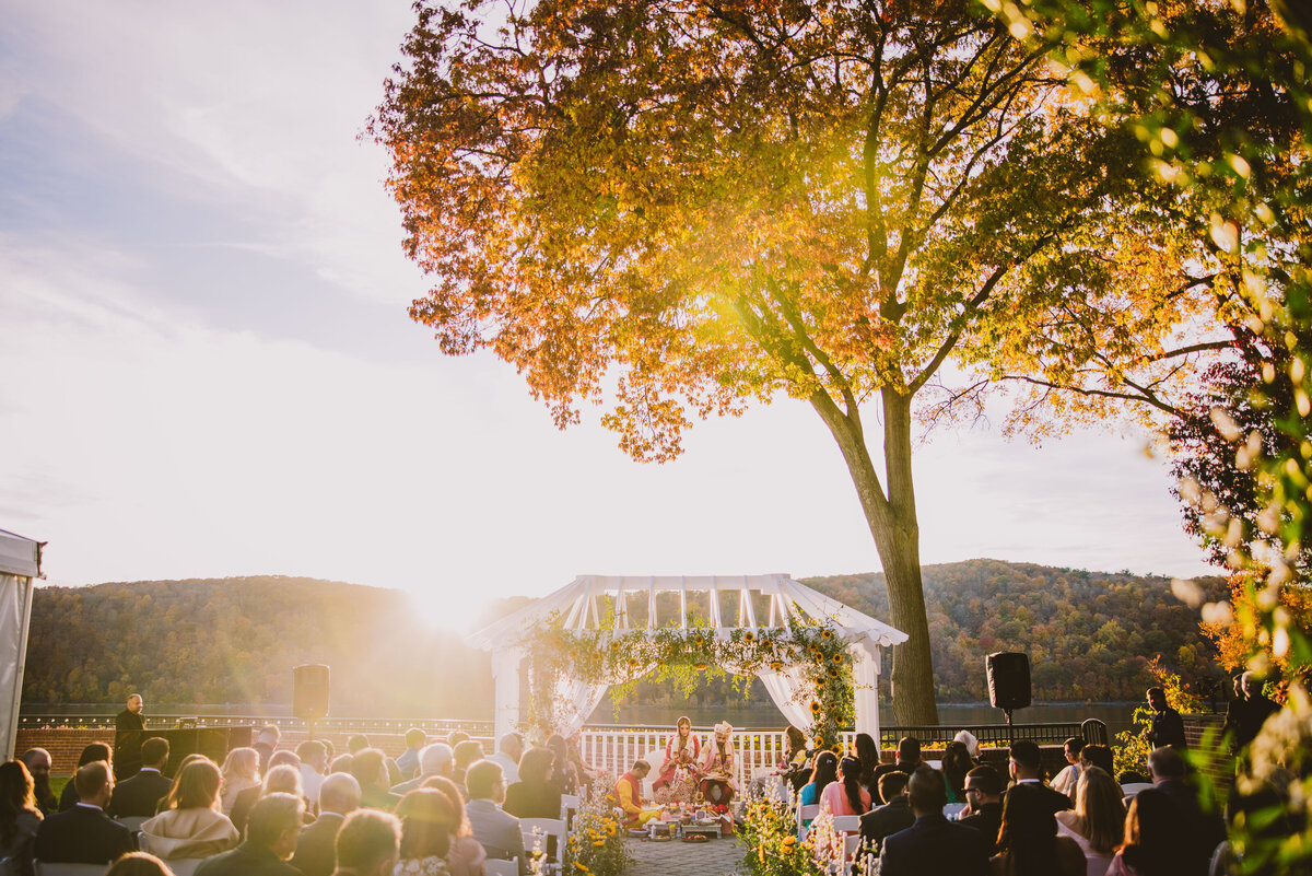 A wide shot of the hindu ceremony taking place at the grandview poughkeepsie in the fall with the sun and fall-colored tree in the background