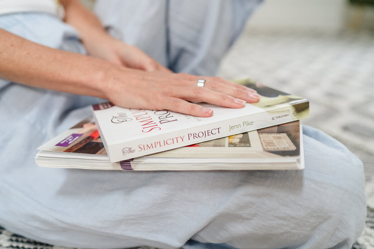 Hand resting on stacked books including The Simplicity Project by Jenn Pike, on crossed legs.