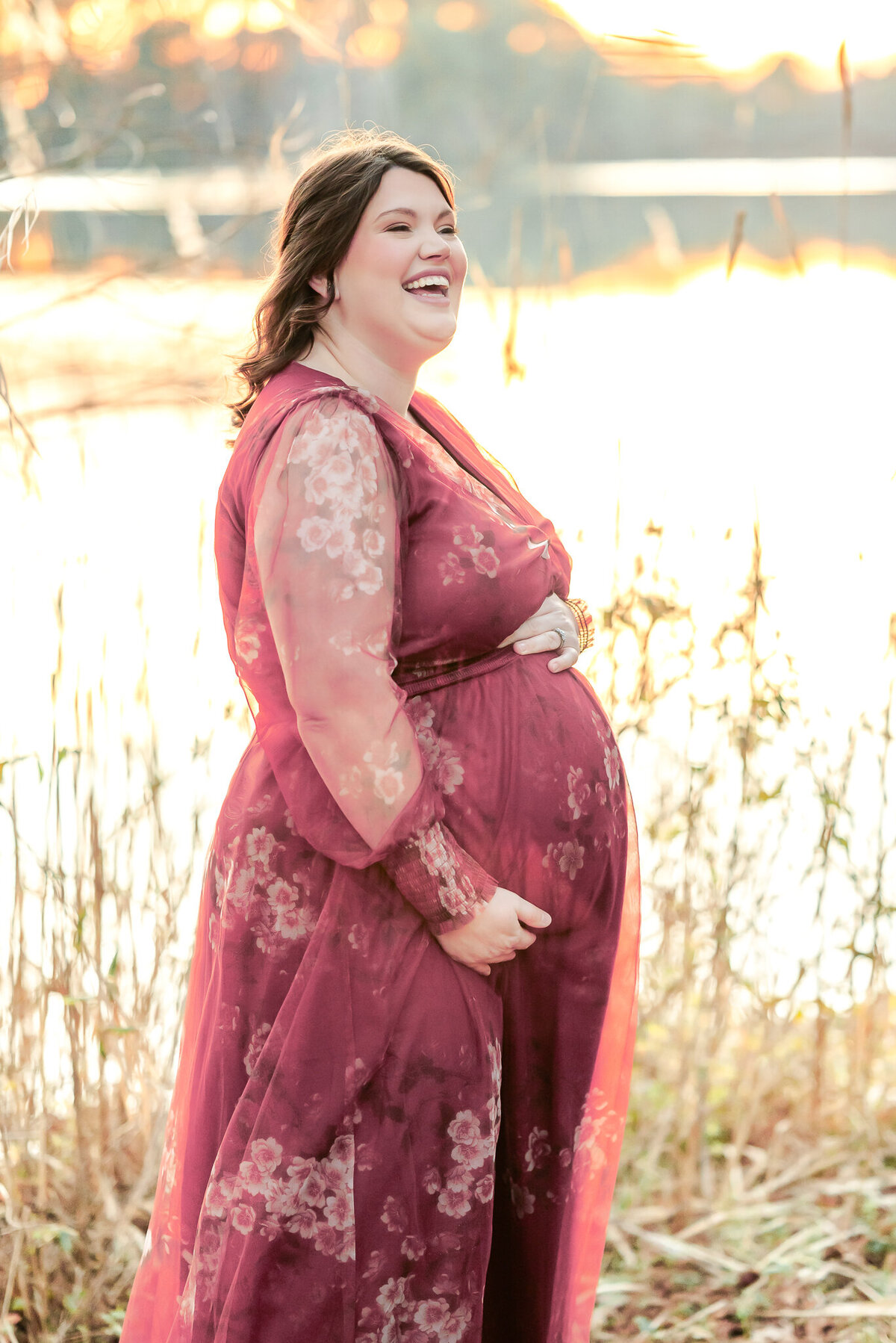 A mama-to-be, wearing a pink floral dress, holds her belly and laughs at the antics of her family.