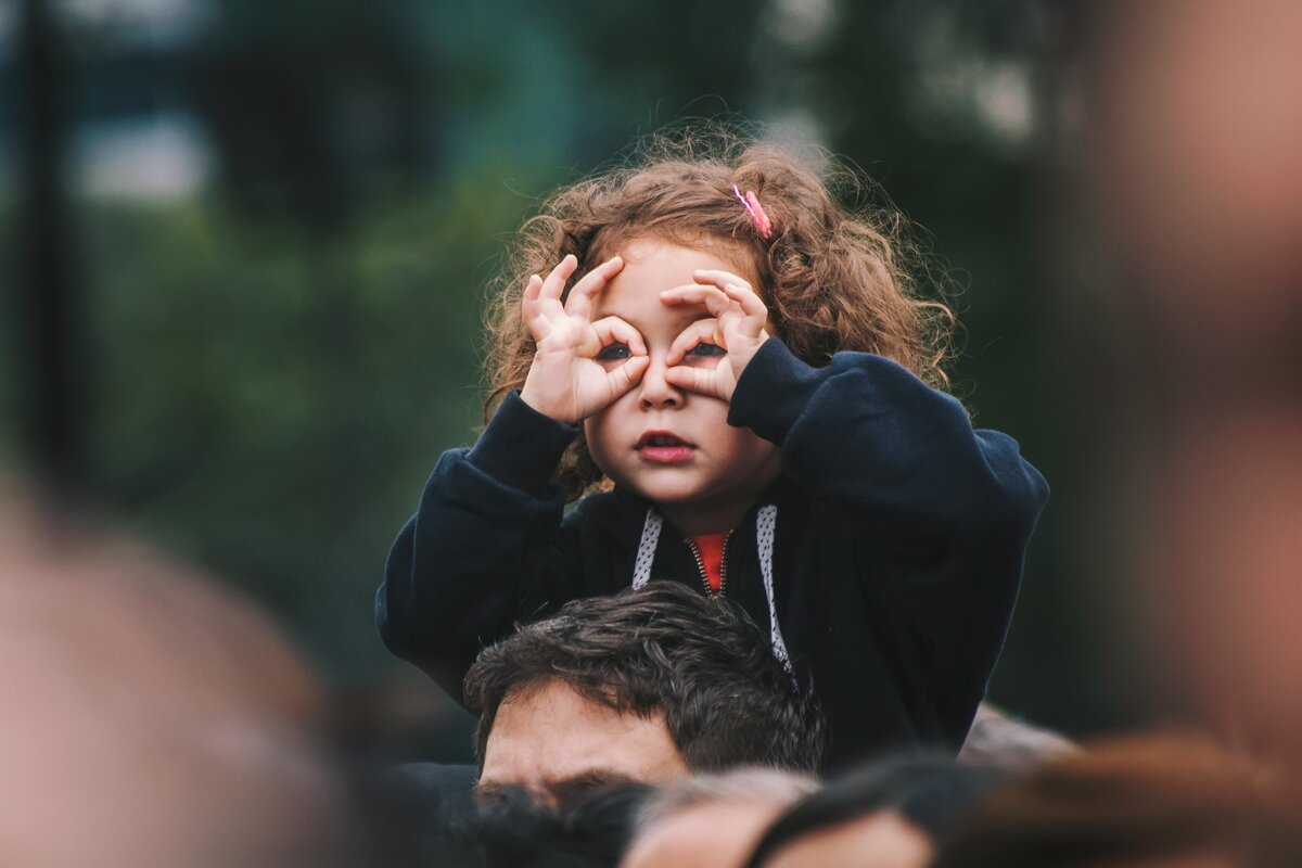 A little girl sits on top of someone's shoulders, holding her hands in front of her eyes like goggles.