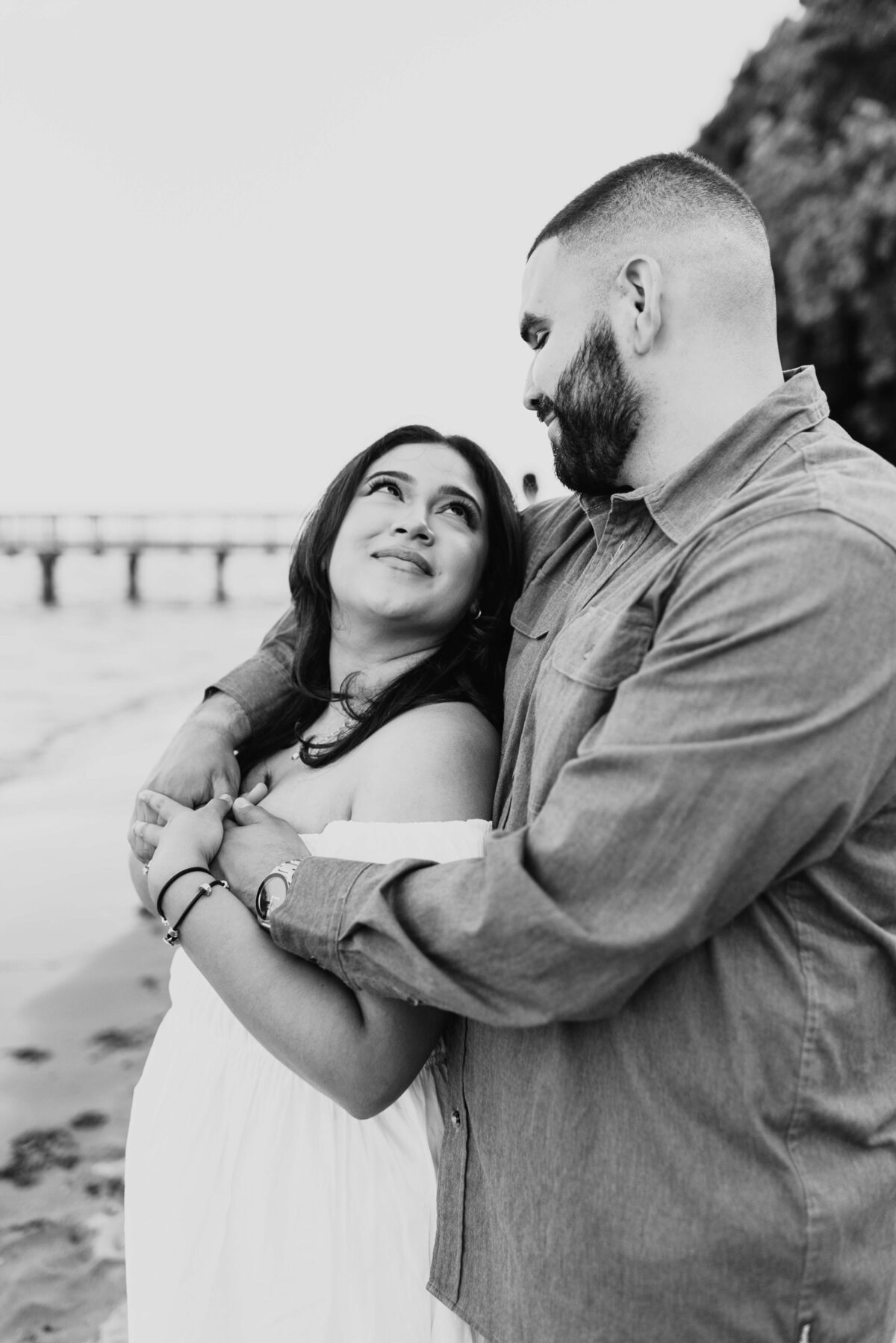black and white engagement photos with woman standing in front of a man while the man holds her hands with his arms around her shoulders while he smiles down at the woman as they stand together on the beach
