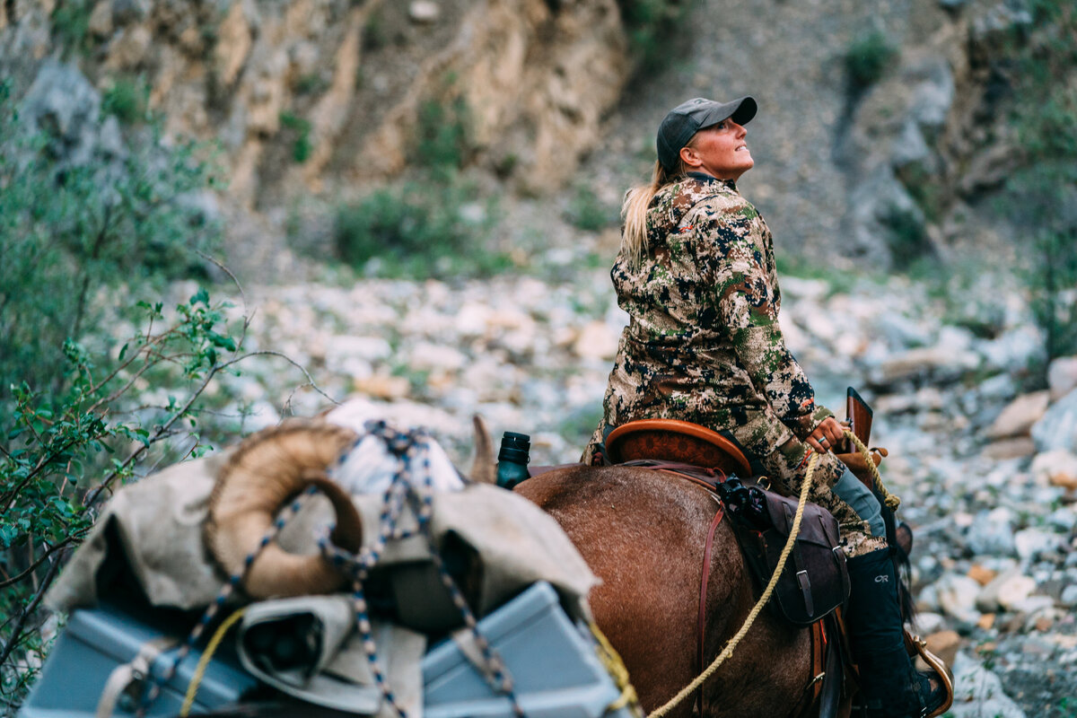 Rachel rides her horse leading a pack string of horses through the mountains gazing up to the right.