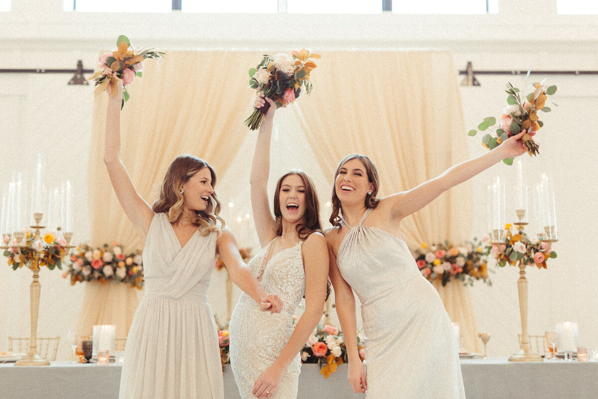 A bride wearing a white wedding gown poses with bridesmaids wearing light gray gowns holding bouquets in the air, smiling.