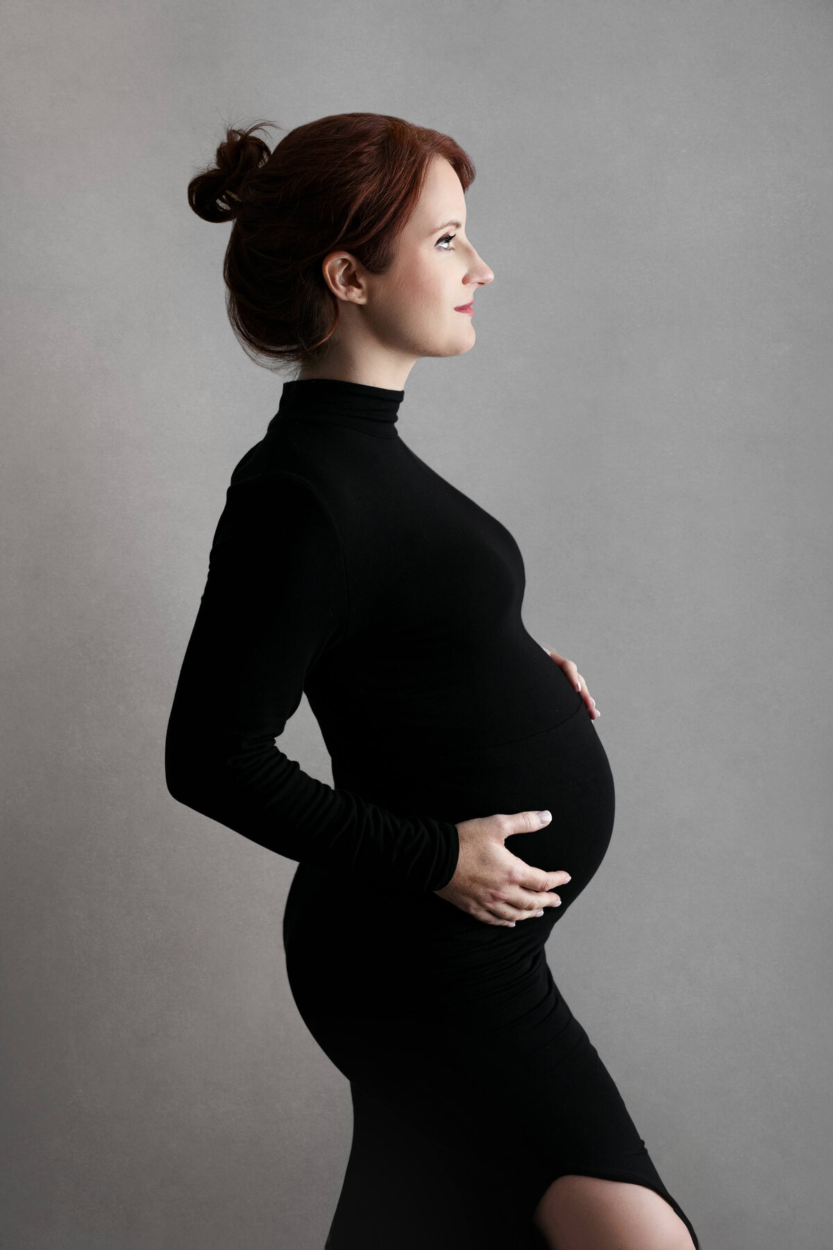 pregnant mother holding her belly wearing a black turtleneck maternity gown on a grey backdrop