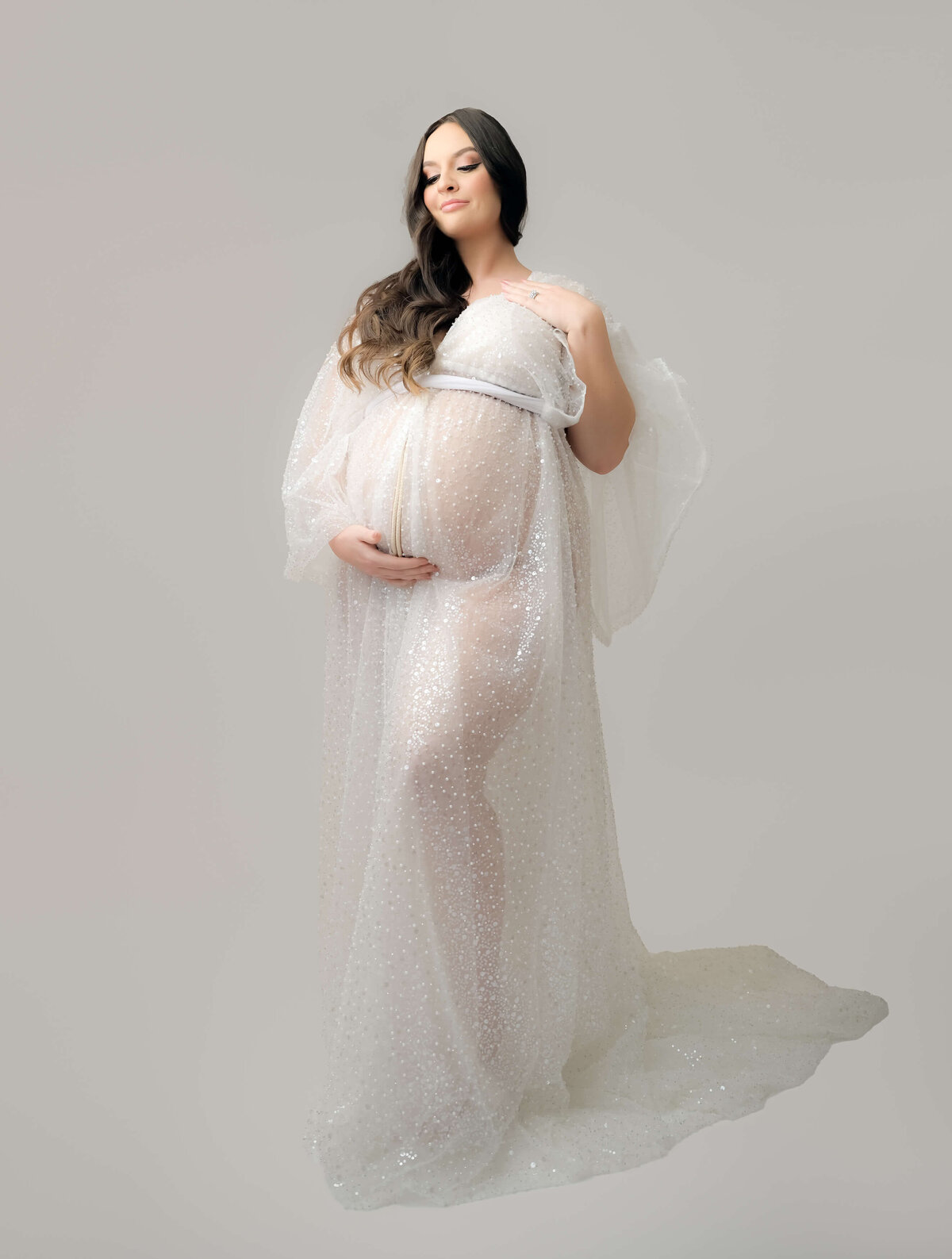 This maternity photo session takes place in Bremerton and is photographed by E. Benecki Photography. The expecting mother is in a white sequin sheer robe. Photographed from a slightly low angle looking up, her long brown hair rests on one shoulder  as she looks down to her belly.