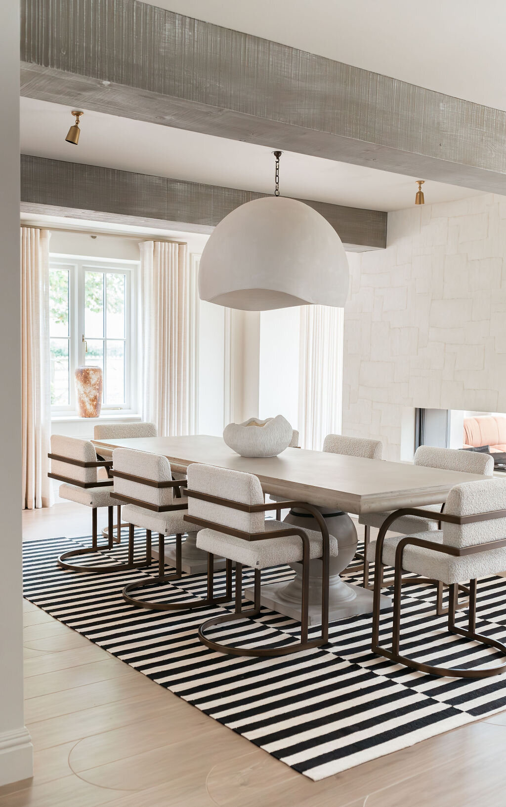 A modern dining room with a large rectangular wooden table surrounded by eight cushioned chairs. A large dome-shaped pendant light hangs above the table, and a black and white striped rug lies underneath. The room features large windows with light-colored curtains.