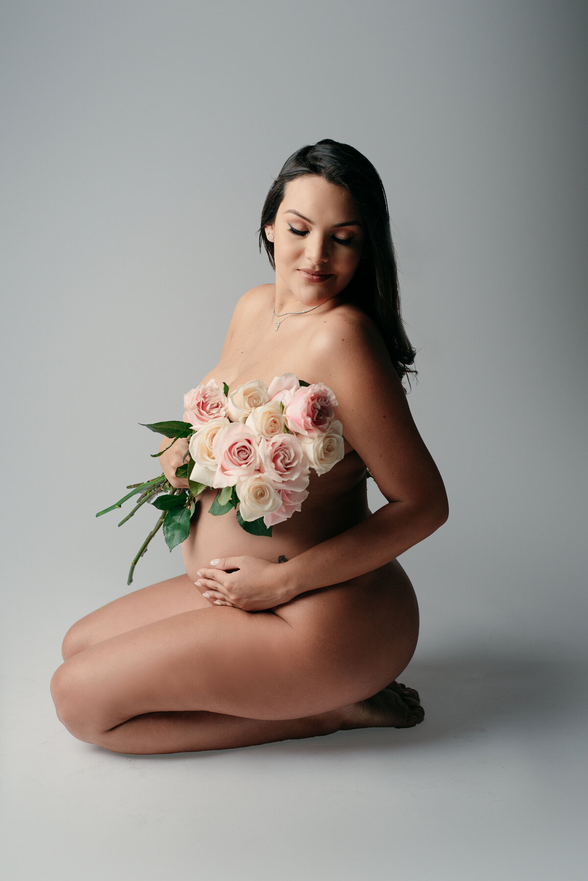 Nude maternity portrait with woman sitting on knees looking over shoulder holding hand on baby bump and holding a bouquet of roses to cover her breast area