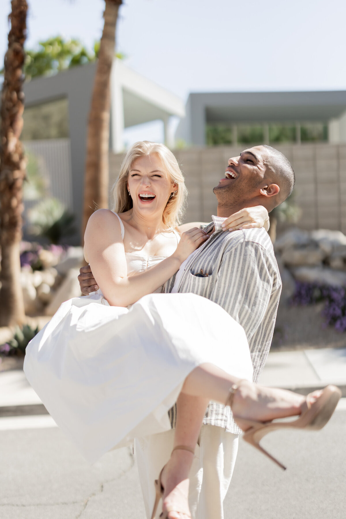 Couple smiling as one lifts the other into the air, captured mid-laugh in bright daylight with modern architecture nearby.