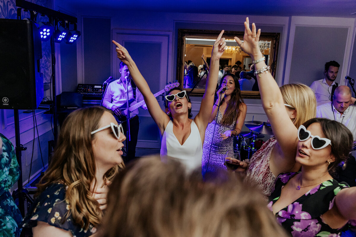 Bride dancing at her wedding reception with her friends, they're all wearing white heart sunglasses
