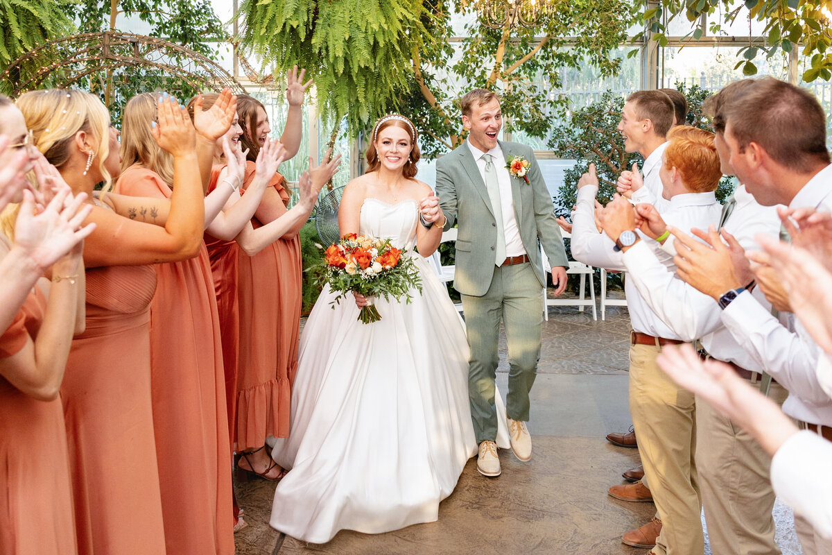 The bride and groom walk hand-in-hand down an aisle of cheering friends and family at Le Jardin in Sandy, Utah. The bride holds a bouquet of orange and white flowers, while the groom beams with excitement