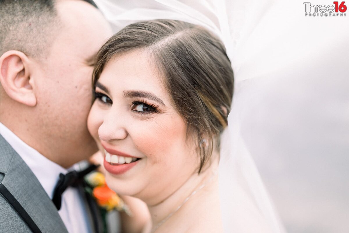 Groom whispers into his Bride's ear as she smiles while they are both under her veil