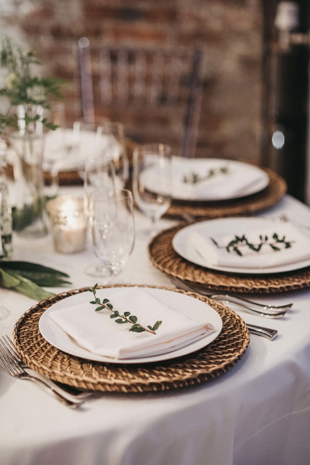 place setting at wedding with rattan charger plates, and napkin with greenery sprig treatment and clear Chivari chairs