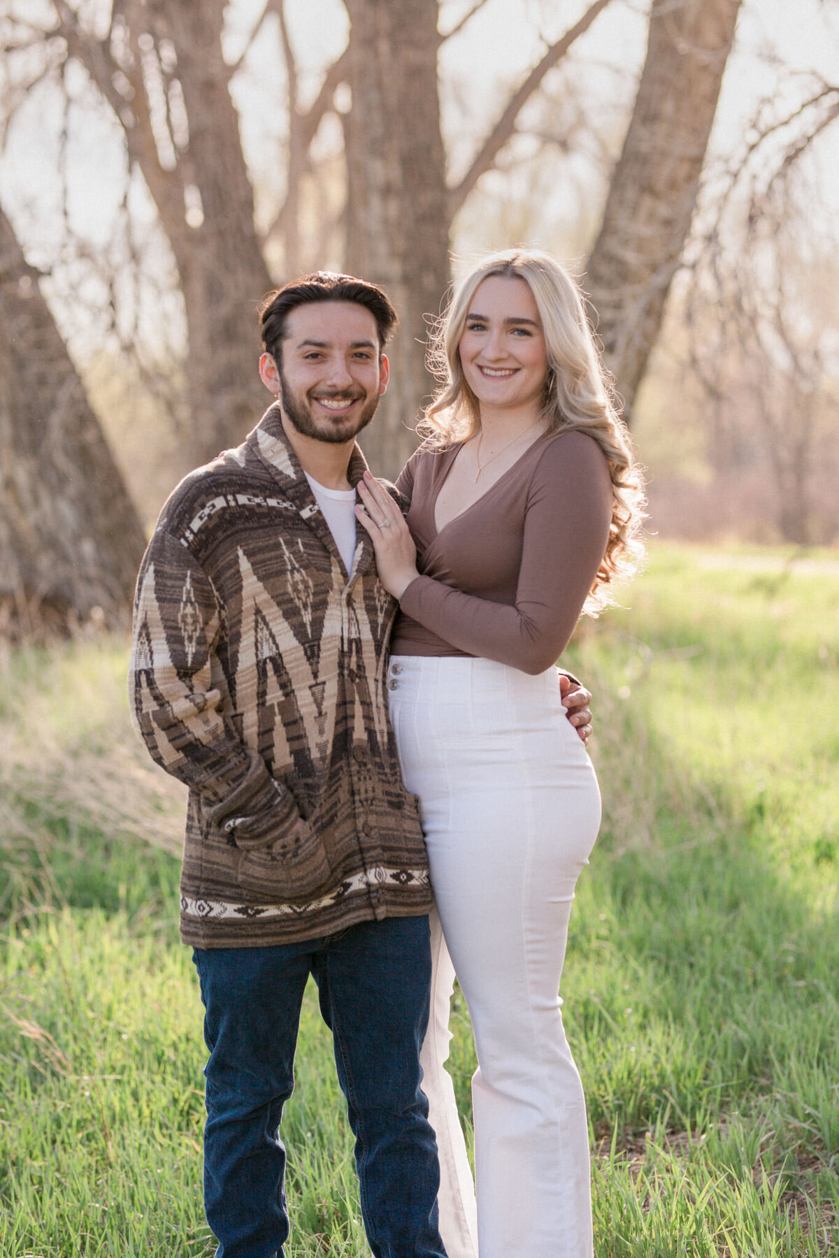 An engaged couple pose with the woman's hand on her man's chest to display their new engagement ring in a wooded area