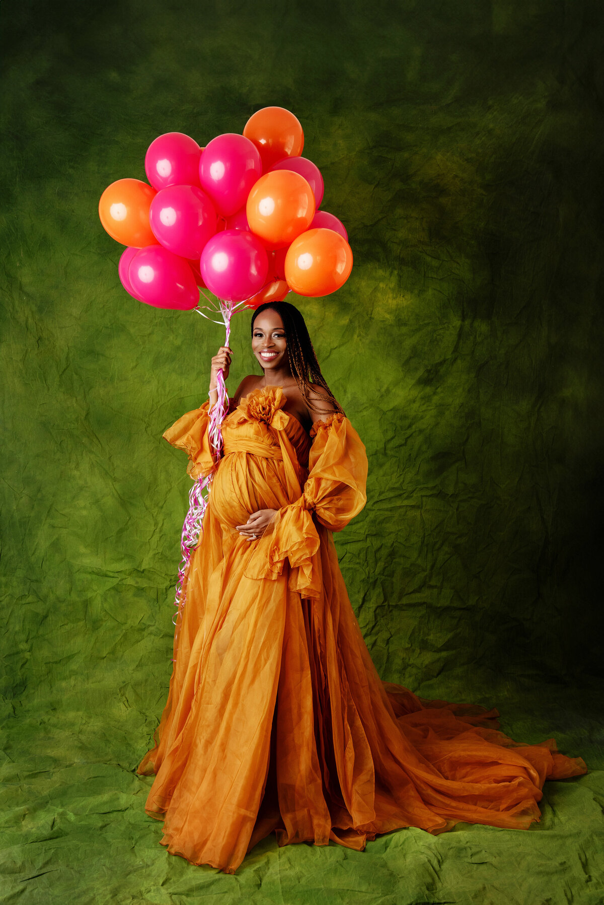 st-louis-maternity-photographer-mom-in-burnt-orange-fluffy-gown-with-orange-and-pink-balloons-on-green-canvas-backdrop