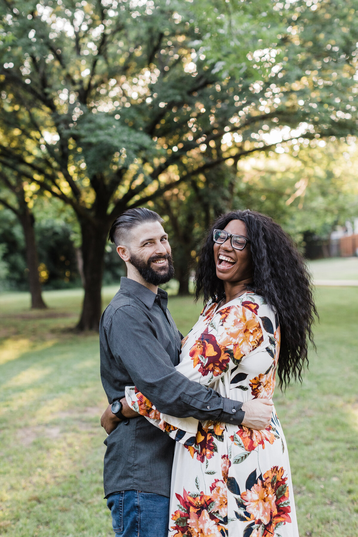 A couple laughing and holding each other close during their outdoor engagement session in Fort Worth, Texas. The woman on the right is wearing a white dress adorned with large, colorful flowers and glasses. The man on the left is wearing a grey dress shirt and jeans.
