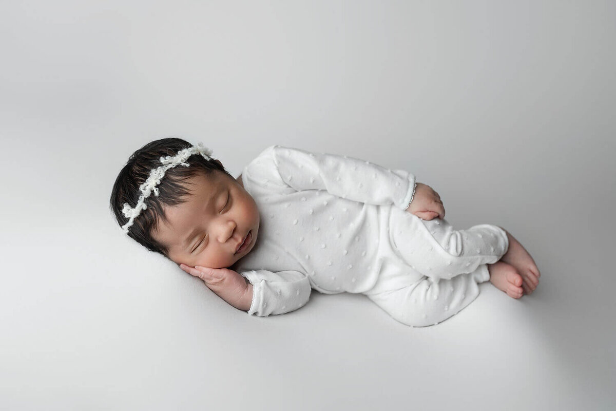 A sleeping newborn baby lays on her side in a white onesie and headband