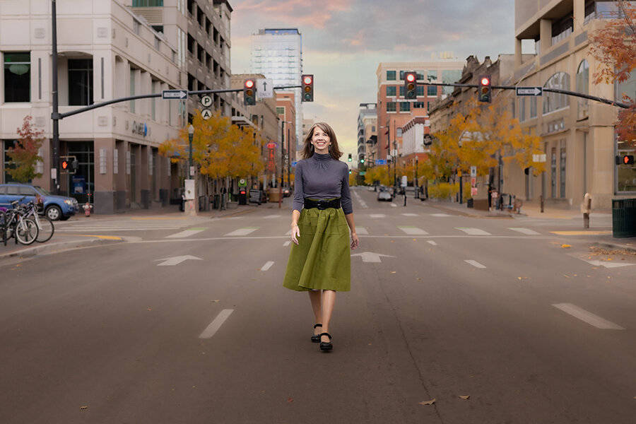 Middle aged woman in grey top and green skirt walking down the middle of a downtown street.