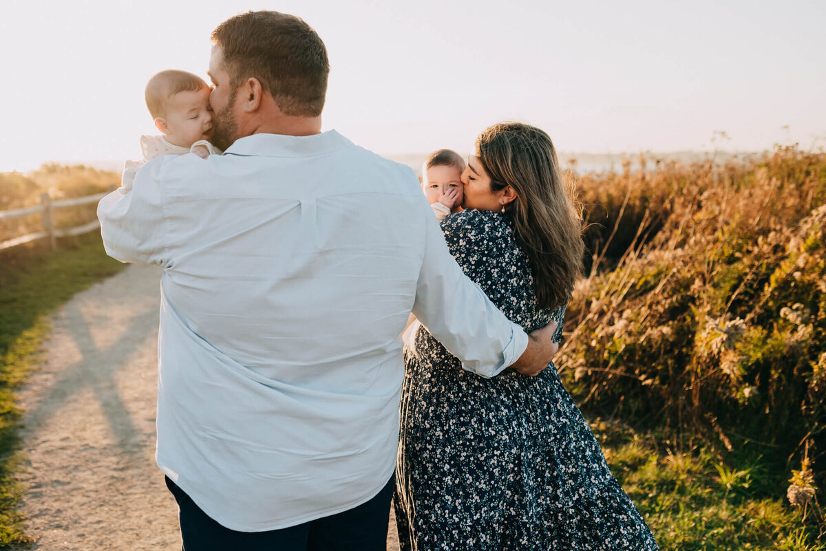 A mother and father hold their twin baby girls while they walk down a path next to the beach at sunset