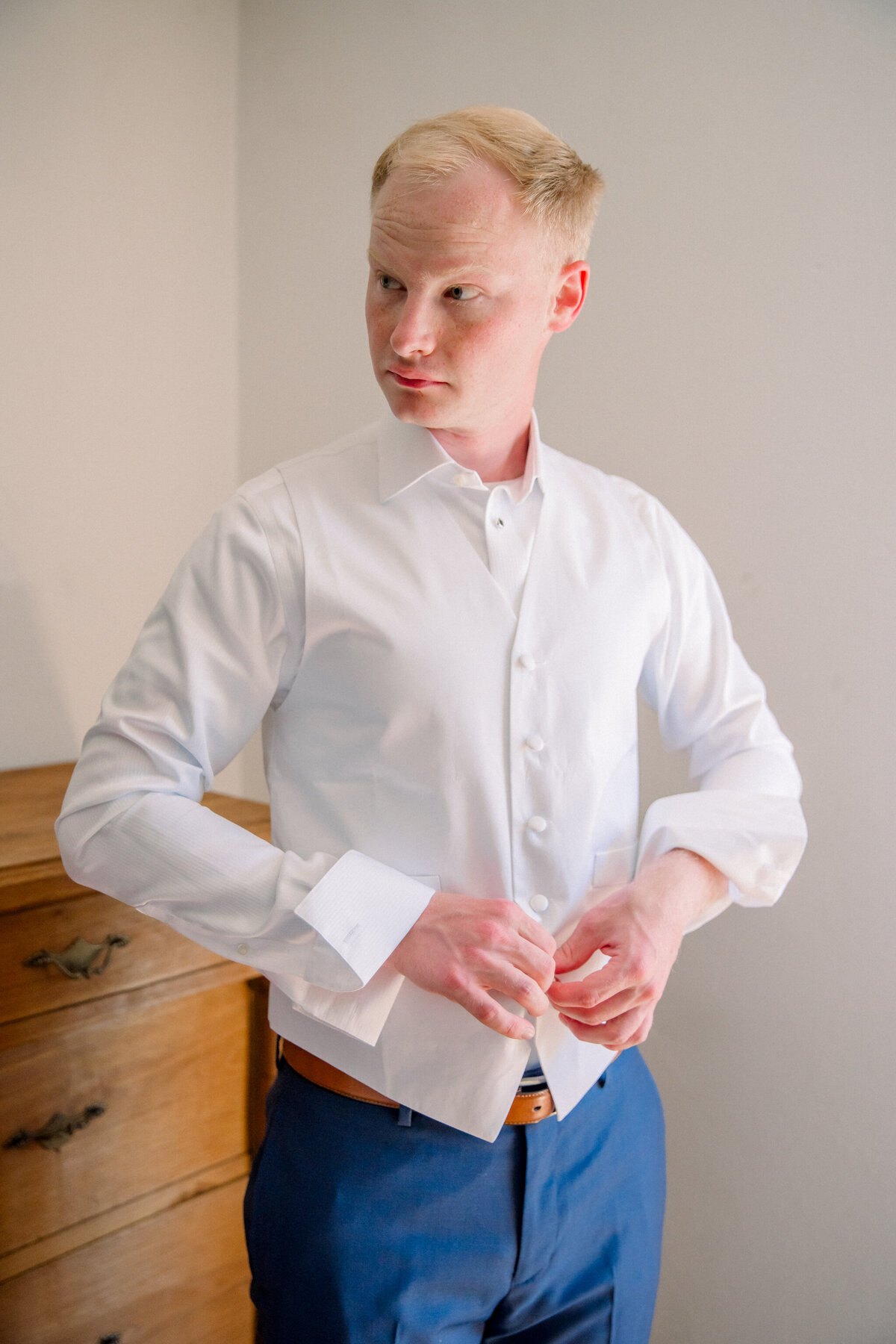 Destination Wedding in Spain | A groom in blue dress pants and a white button-up shirt buttons his white vest while getting ready for his wedding in Spain.