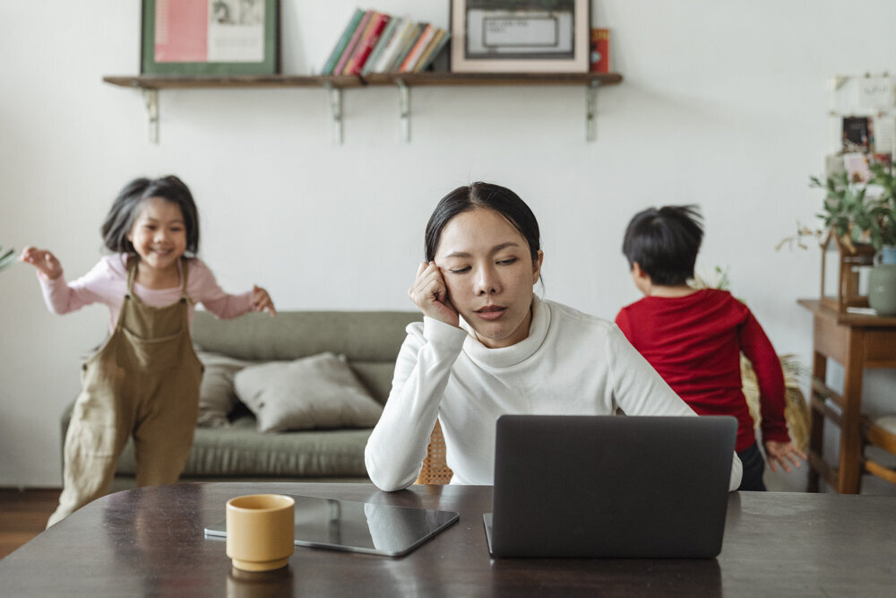 An Asian woman wearing a white turtleneck rests her face in her right hand, staring down at a laptop with an exasperated expression. Two young children run in the background of the image.