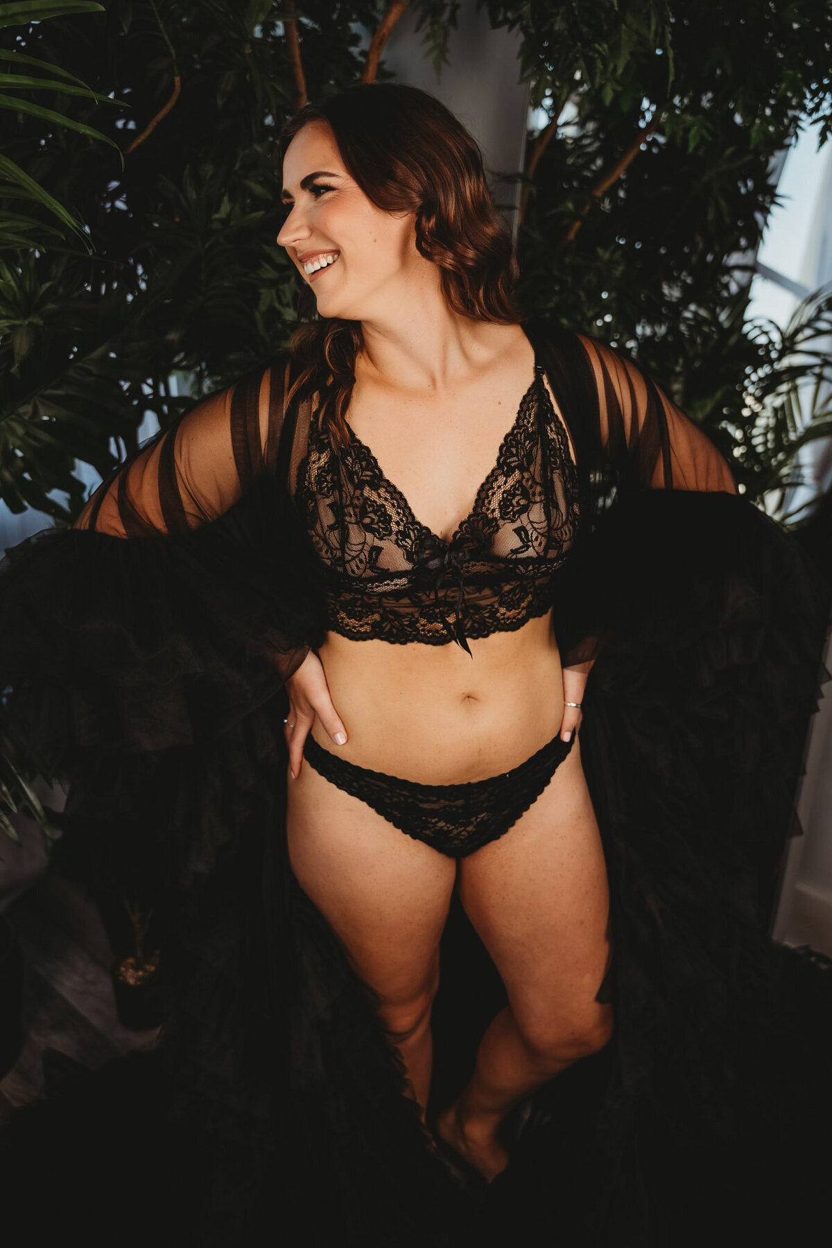 smiling woman wearing black two piece lace lingerie and a giant black tulle robe, while laughing away from the camera