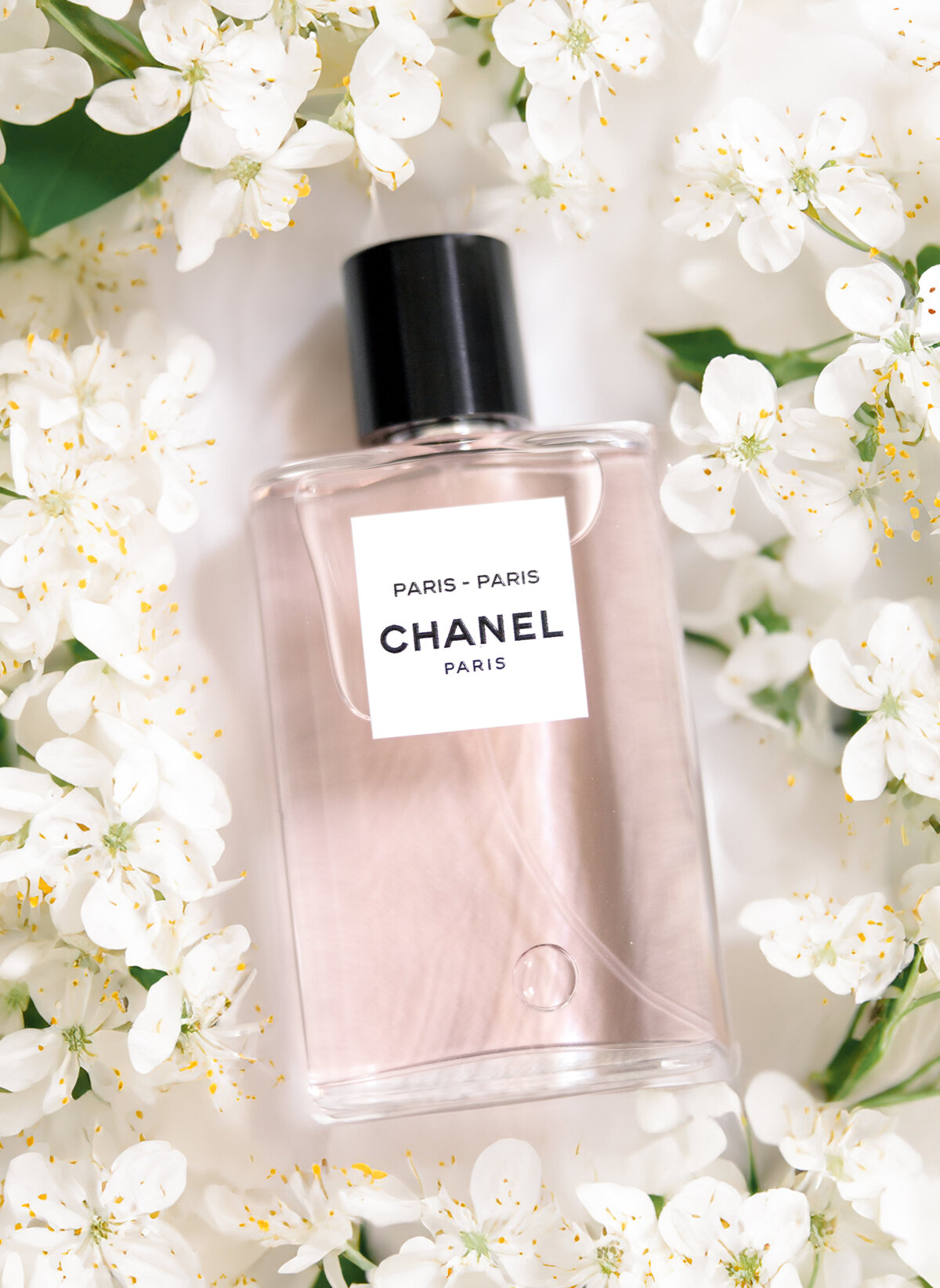 A bottle of Chanel Paris perfume lies on a bed of white flowers. The bottle is clear with a black cap and features a simple, elegant design. The floral background complements the perfumes sophisticated aesthetic.
