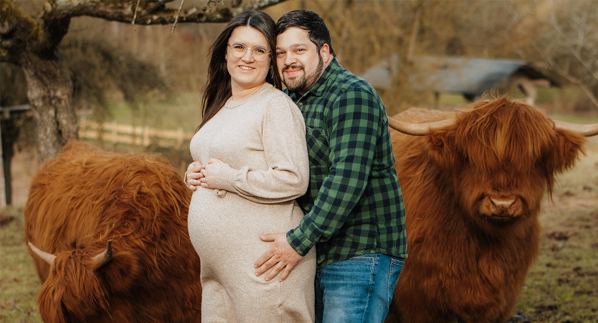Ein Ehepaar steht auf der Weide, umfasst den Babybauch und neben ihnen stehen zwei Highland Rinder