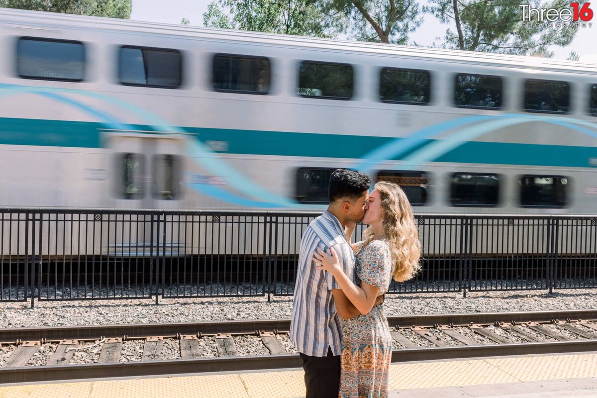 Engaged couple share a kiss next to the train tracks while the train sits behind them