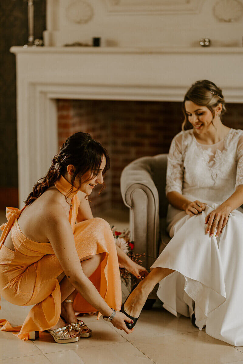 Demoiselle d'honneur vêtue d'une longue robe couleur abricot enfilant l'escarpin de la mariée assise dans un fauteuil devant une cheminée. Shooting photo mariage.