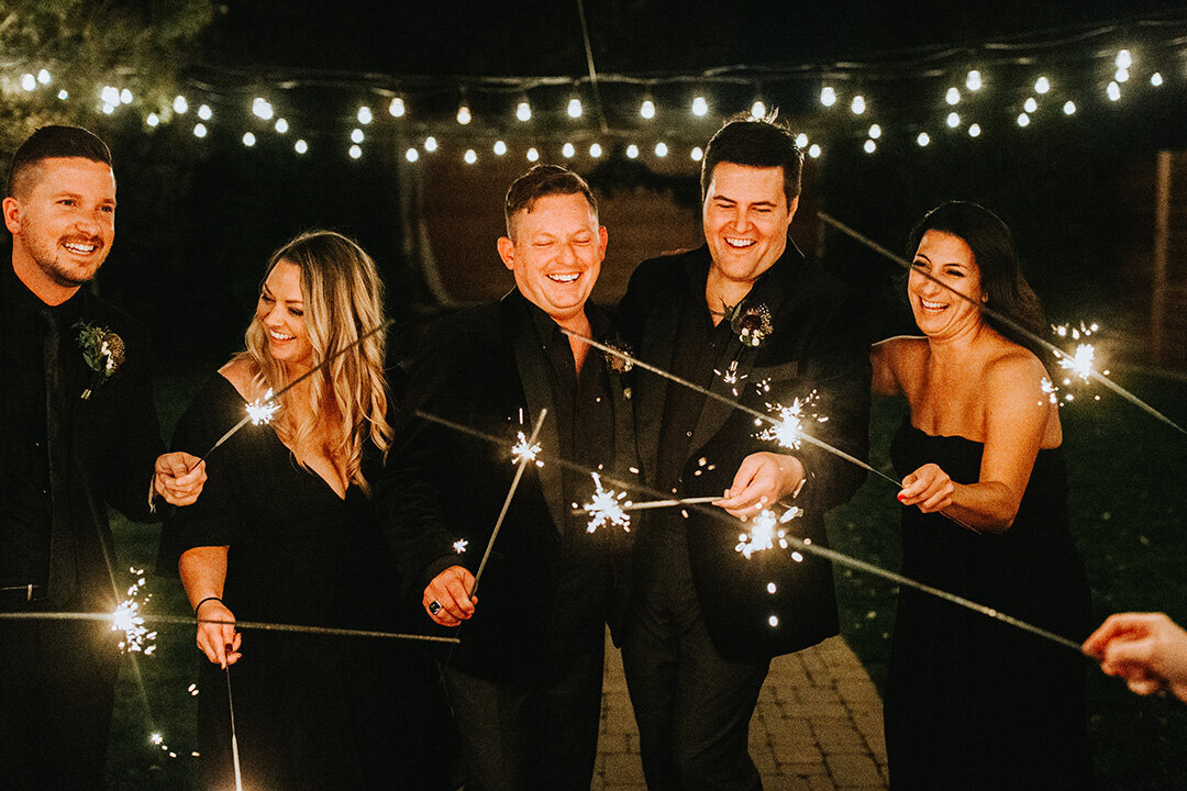 Two grooms wearing black tuxedos hold sparklers in their hands at night with friends at the wedding reception.