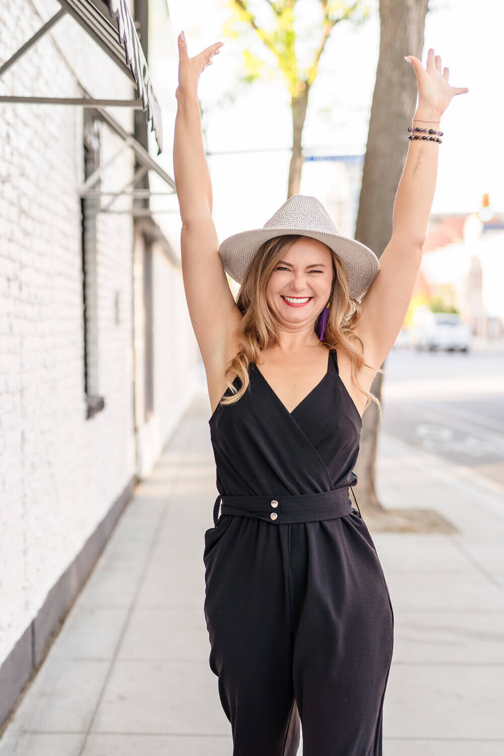 Entrepreneur walking down sunny city sidewalk in black dress, smiling toward the camera with hands raised in celebration.